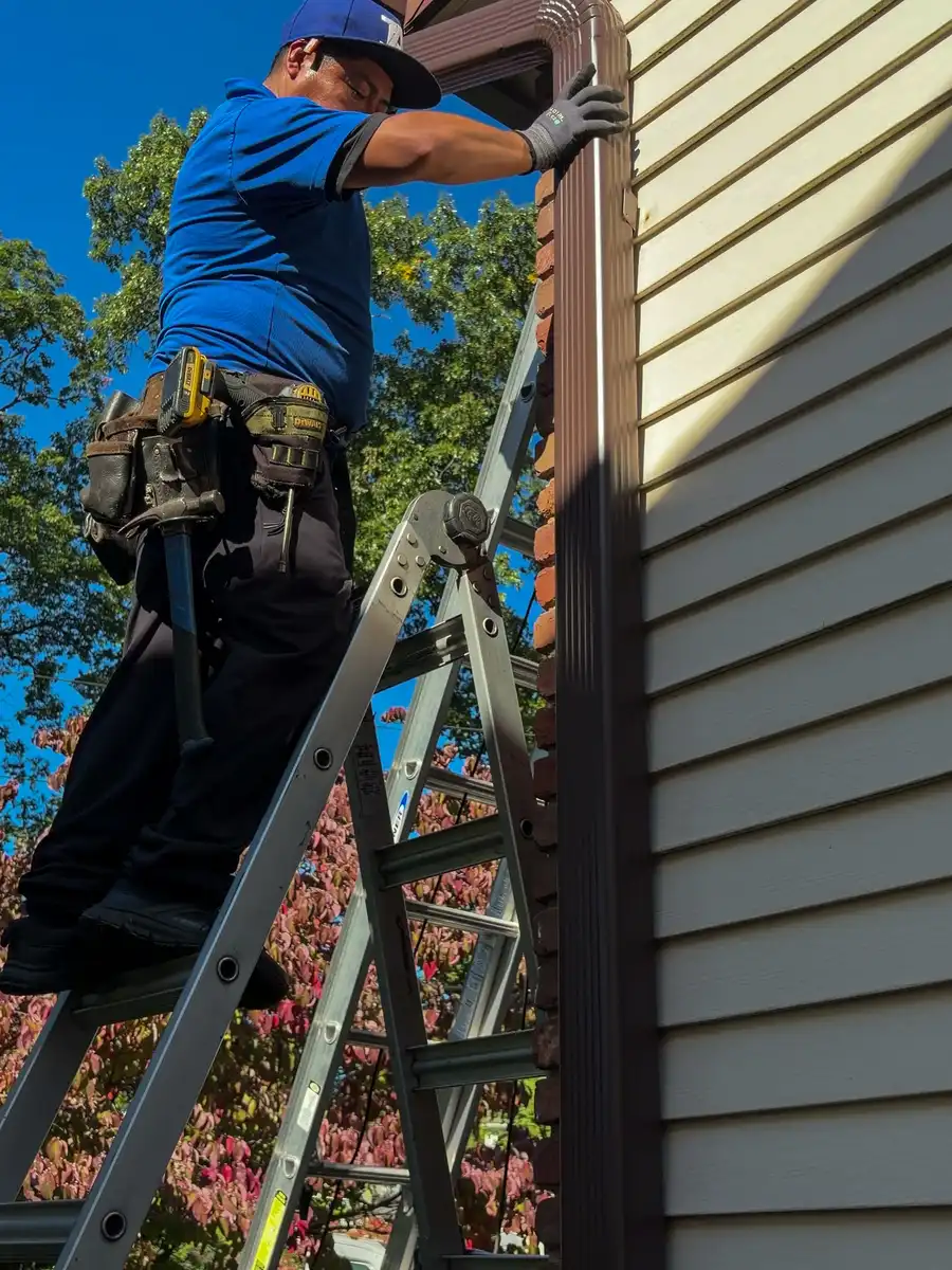 Tav crew member in a blue Tav shirt on a ladder fastening a new brown gutter and downspout at the roof edge, blue sky